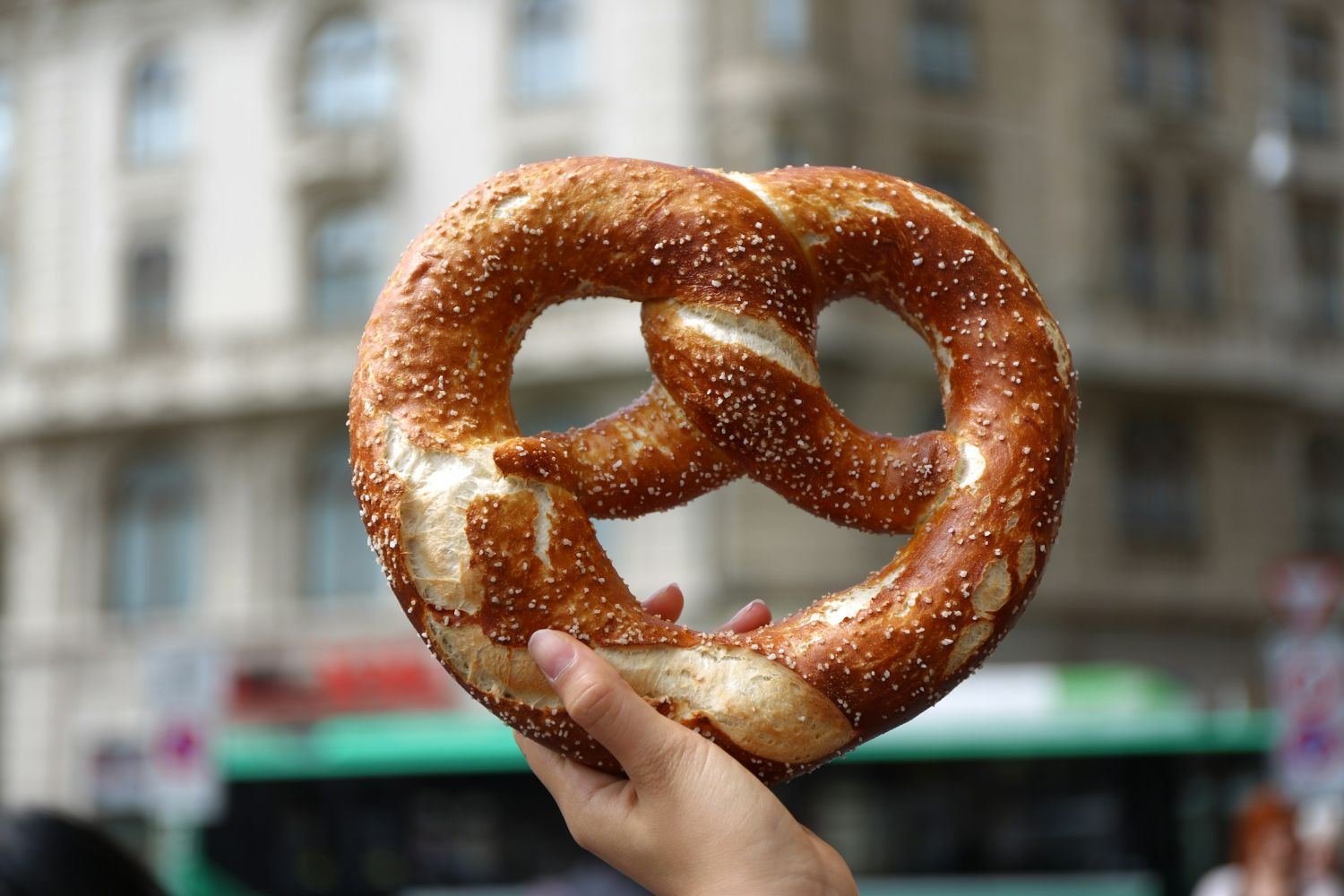 Une main tient un grand bretzel doré devant une rue animée